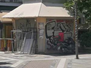 Παραδοσιακα ελληνικά περίπτερα σε αστική γωνία, με προϊόντα και περιοδικά, σε φόντο σύγχρονης πόλης.